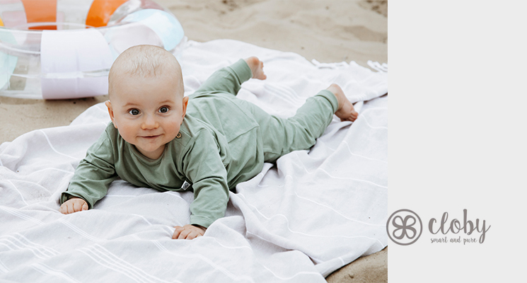 Kind mit grünem Longsleve und Hose liegt auf einer Decke am Strand. Marke cloby