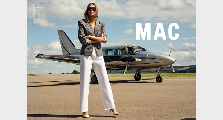 Frau stehend vor Kleinflugzeug mit grauem Blazer und weißer Hose von Mac