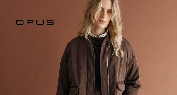 Frau stehend mit gestreifter Bluse, braunem Pullover, brauner gesteppter Jacke und blauer Jeans mit Einkaufstüte von Opus