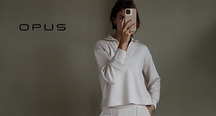 Stehende Frau mit weißem Pullover und weißer Hose mit Handy in der Hand von Opus