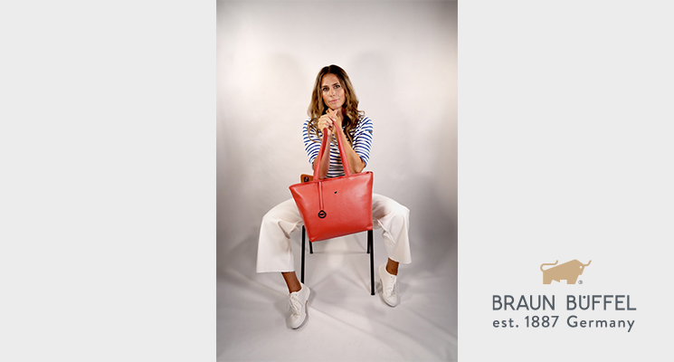 Frau sitzend auf Stuhl in gestreiftem Shirt und weißer Hose, welche eine orangene Tasche von Braun Büffel in den Händen hält