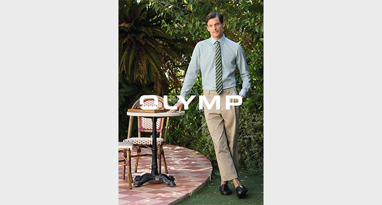 Mann an Tisch lehnend in grün weiß gestreiftem Hemd mit grün weiß gestreifter Krawatte und beiger Hose von Olymo