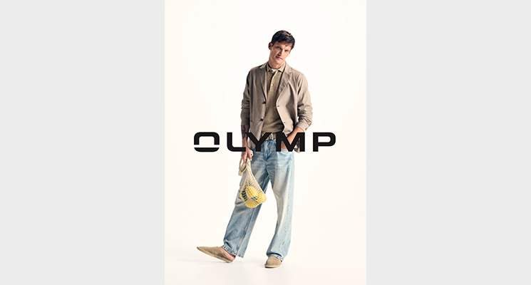 Stehender Mann mit beigem Sakko und dunkelbeigem Hemd, blauer Jeans und Zitronennetz in der Hand von Olymp