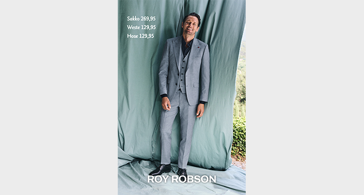 Mann in hellblauem Anzug und schwarzem Hemd von Roy Robsen vor Vorhang stehend