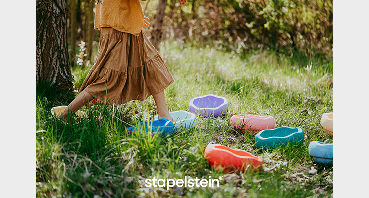 Bunte Stapelsteine auf Wiese verteilt und Mädchen läuft von Stein zu Stein, welche sie zu einem Weg gelegt hat
