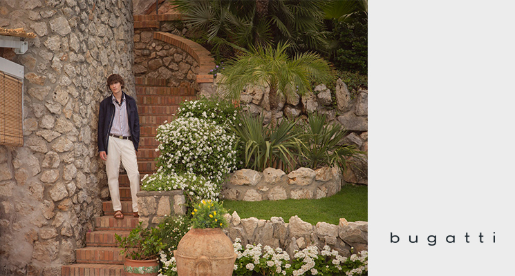 Mann in blühendem Garten mit blauer Jacke, gestreiftem Hemd, beiger Hose und braunen Sandalen