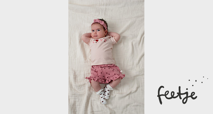 Baby auf Bett liegend mit beigem T-Shirt und rosefarbenen kurzen Hose von Feetje