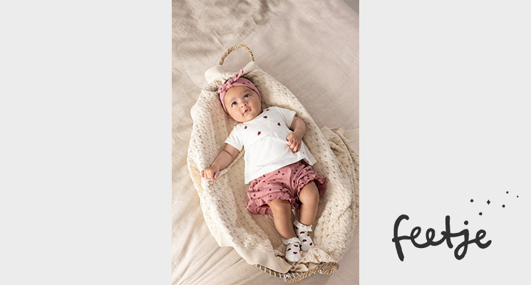 Baby in weißem T-Shirt mit Muster und rose farbener kurzen Hose, in Körbchen liegend von Feetje