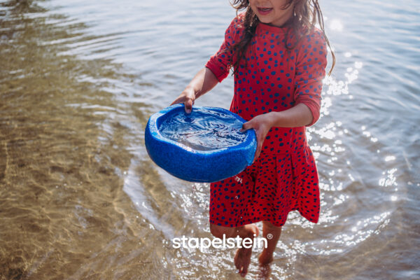 blauer Stapelstein als Schüssel zum Spielen verwendbar. Hier sieht man ein Mädchen am Wasser.