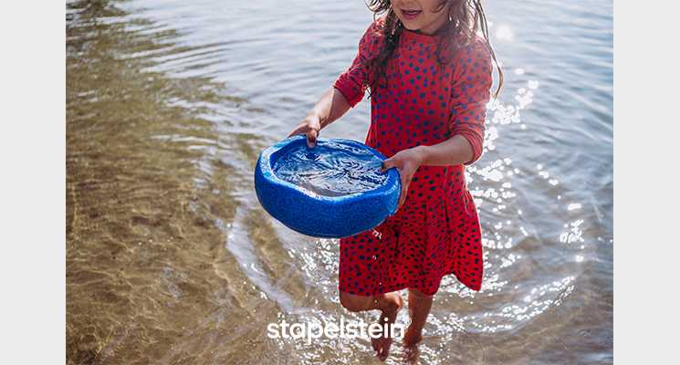 blauer Stapelstein als Schüssel zum Spielen verwendbar. Hier sieht man ein Mädchen am Wasser.