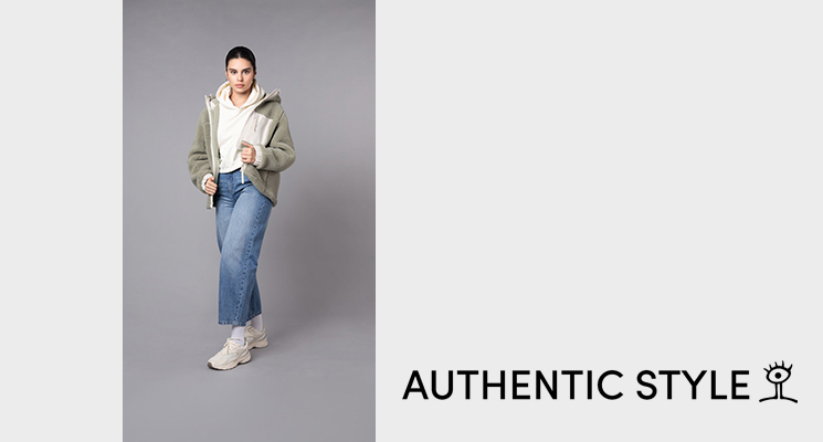 Stehende Frau mit weißem Kapuzenshirt, darüber eine weißkhakifarbene Teddy Felljacke dazu eine blaue Jeans von Authentic Style