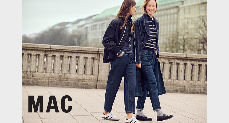 2 stehende Frauen: eine trägt blauen langen Mantel mit blau weiß gestreiftem Oberteil und blauer Jeans, die andere Frau trägt schwarze Reißverschlussjacke und blaues Jeansoberteil mit Knöpfen und blaue Jeans von Mac