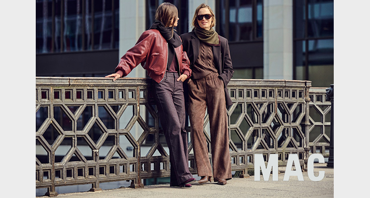 2 stehende Frauen eine davon trägt pflaumefarbenen Mantel mit dunkelbraunem Strickoberteil und braune Stoffhose. Die andere Dame trägt braune Lederimitat Jacke Mit pflaumefarbenem Oberteil und pflaumfarbener Stoffhose von Mac