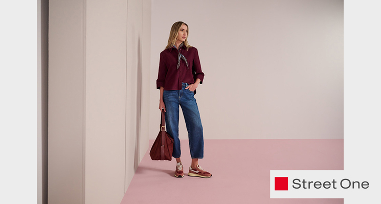 Stehende Frau mit bordeauxfarbenem Jäckchen und Shirt sowie blauer Jeans und Tasche von Street One
