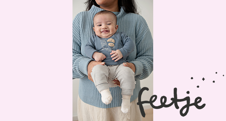 Frau hat Baby auf dem Arm welches einen blauen Pullover mit Motiv und weiße Hose mit beigen Streifen von Feetje trägt