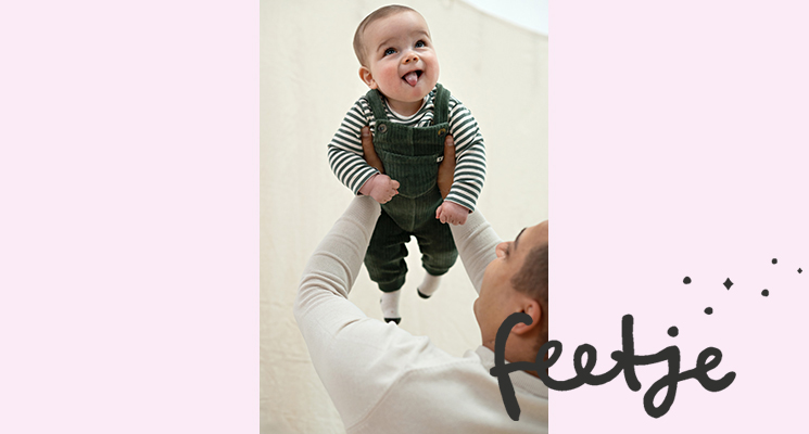 Man hält Babyjunge hoch und dieser trägt gestreiftes Langarmshirt in Khaki weiß und jeansfarbene Latzhose von Feetje
