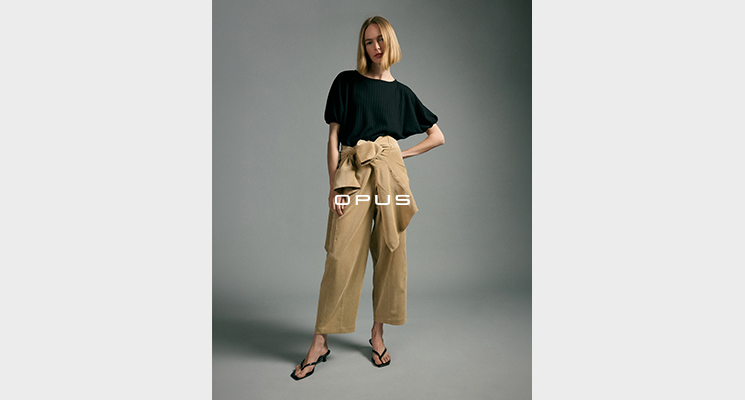Stehende Frau in schwarzem Shirt, hellbrauner Wideleg Stoffhose mit umwickelter hellbrauner Jacke von Opus