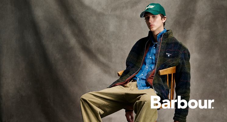 Sitzender Mann mit khakifarbener Jacke blauem Hemd und beiger Hose sowie Cap von Barbour