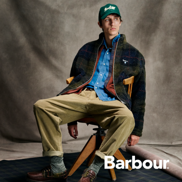 Sitzender Mann mit khakifarbener Jacke blauem Hemd und beiger Hose sowie Cap von Barbour