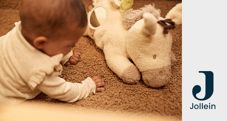 Krabbelndes Baby auf braunem Teppich mit weißem Rüschchenoberteil mit davorliegendem Spielkuscheltier von Jollein
