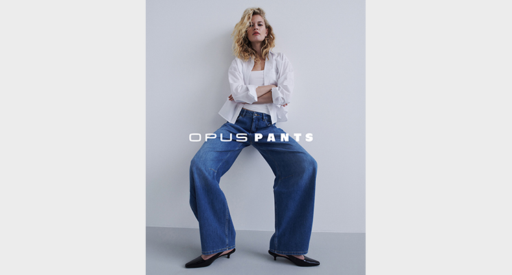 An der Wand lehnende Frau die eine weiße Bluse und Top von OPUS und blaue Jeans von OPUS PANTS trägt
