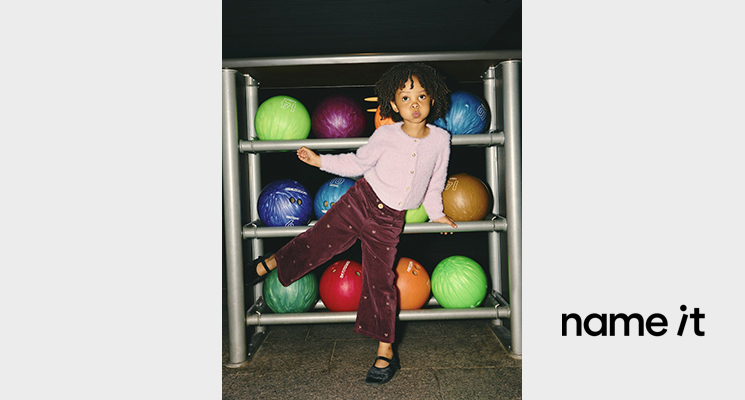 Mädchen in rosa Strickjacke und braunem Hose mit kleinen Motiven vor Ballregal von Name it