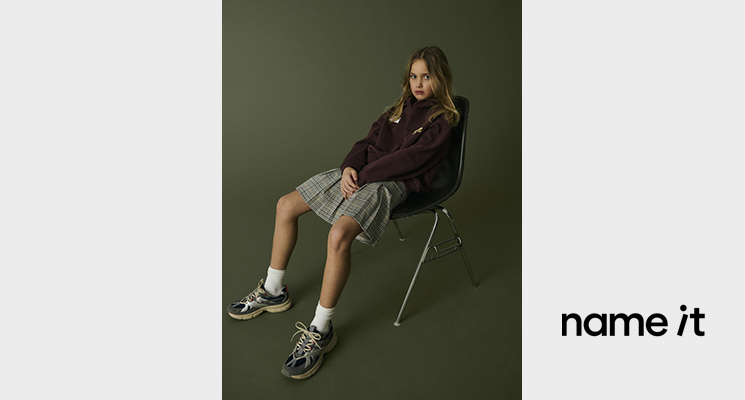 Sitzendes Mädchen auf Stuhl mit braunem Pullover, kariertem Rock von Name it
