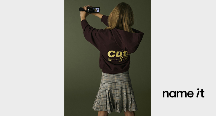 Mädchen mit braunem Pullover mit Backprint Schriftzug in gelb und kariertem Rock von Name it