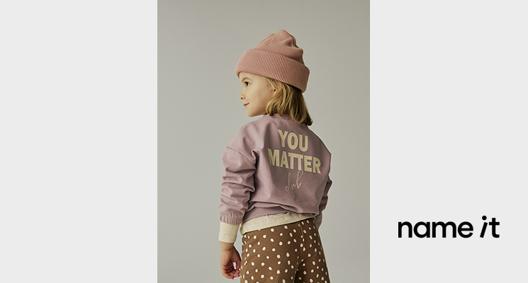Mädchen mit fliederfarbenem Oberteil mit Backprint Schrift sowie braun weiß gepunkteter Hose und rosa Mütze von Name it