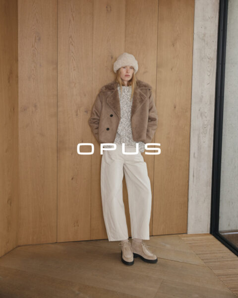 Stehende Frau mit brauner Jacke inklusive braunem Innenfell und grau weiß meliertem Pullover und weißer Hose mit naturfarbener Mütze von Opus