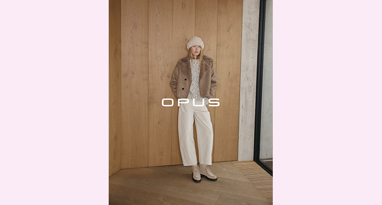 Stehende Frau mit brauner Jacke inklusive braunem Innenfell und grau weiß meliertem Pullover und weißer Hose mit naturfarbener Mütze von Opus
